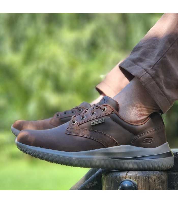 Zapatos Skechers Glavine impermeables marrón en perfil lateral, puestos con pantalones marrones en un parque.